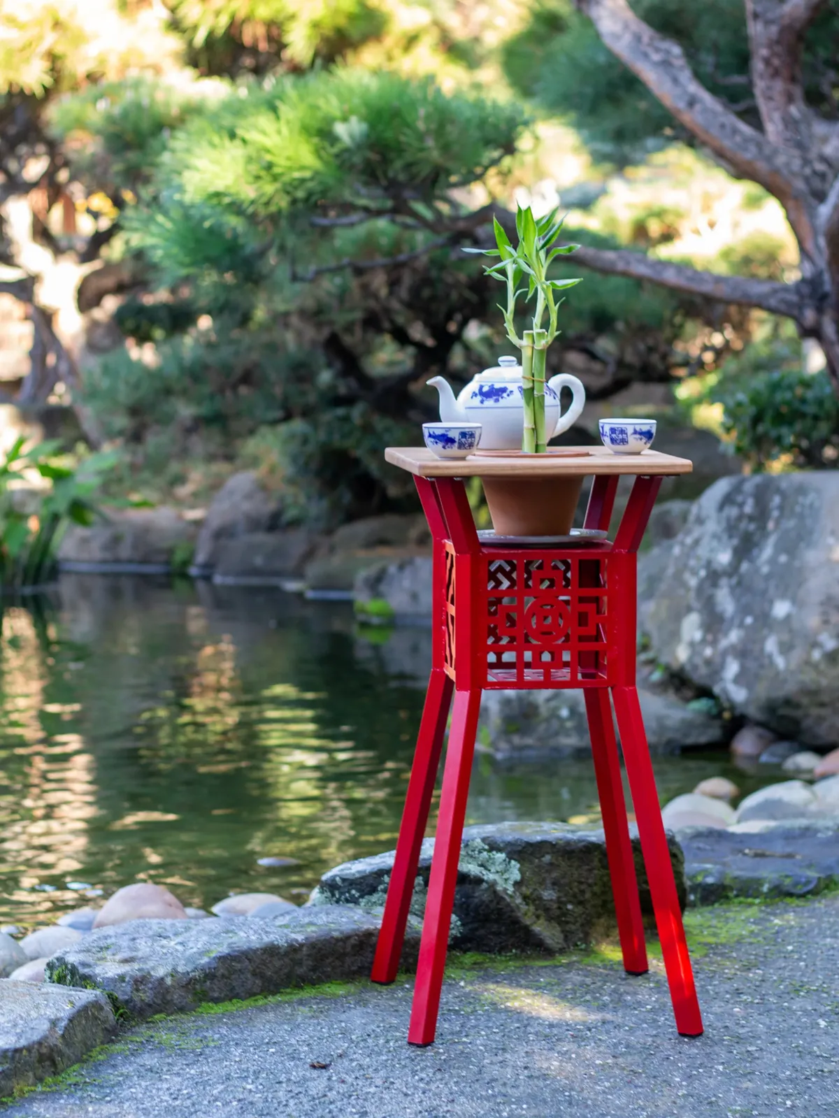 Red steel and wood planter table holding a plant and tea set beside a garden pond