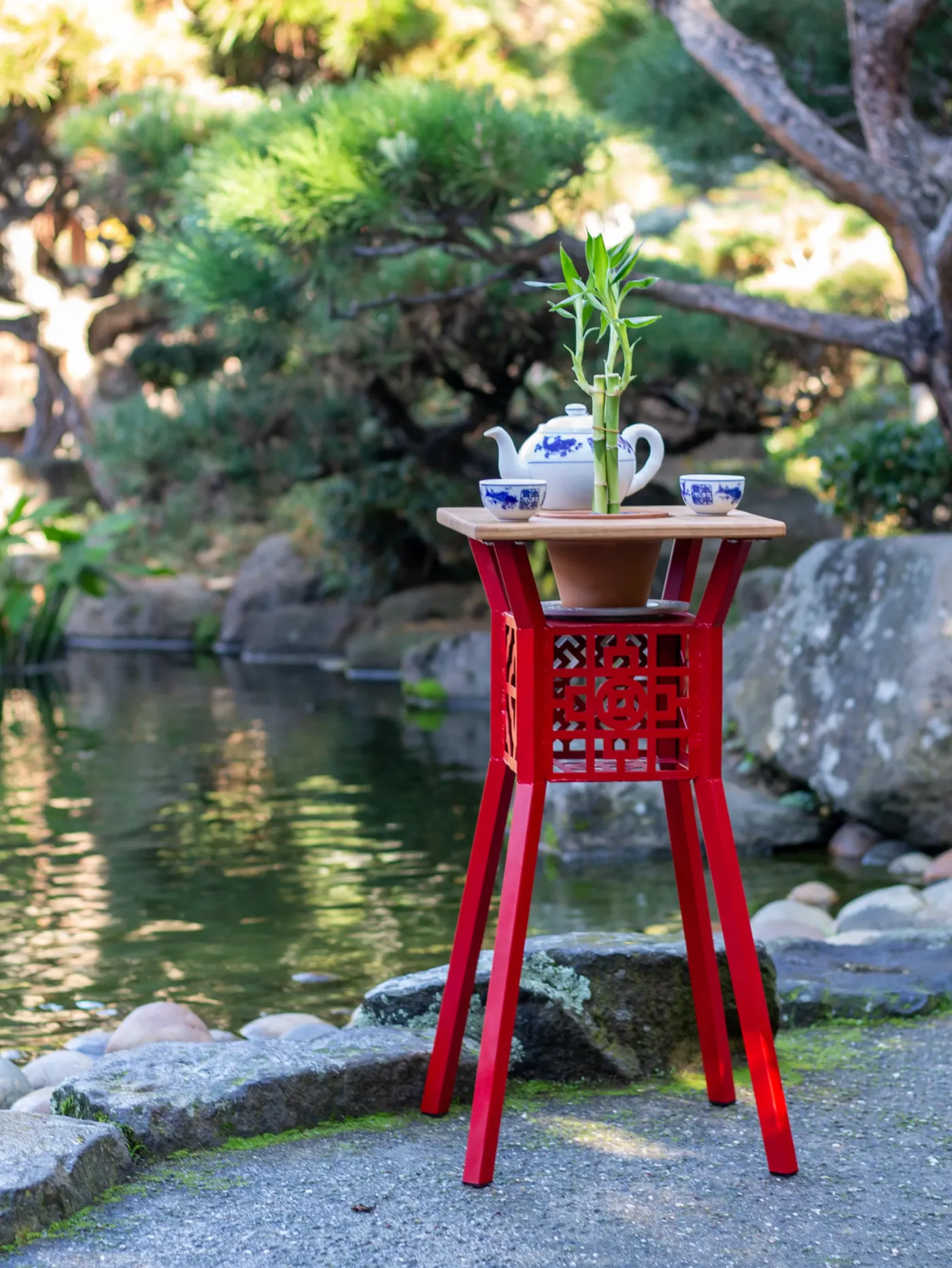 Red steel and wood planter table holding a plant and tea set beside a garden pond